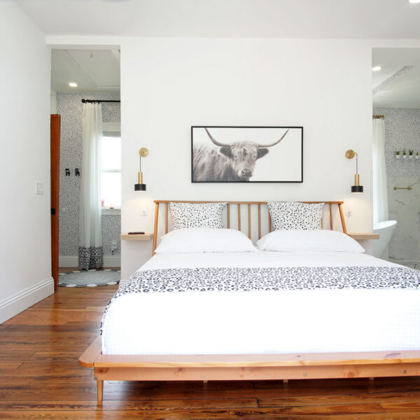 08-shotgun-house-primary-bedroom-king-bed-wood-floors King bed framed by brass sconces with a doorway at right revealing the freestanding soaking tub beyond