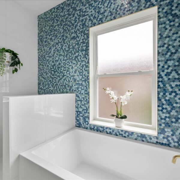 Detail of a white soaking tub set against blue mosaic tile beneath a frosted double hung window