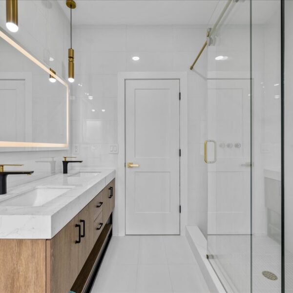 Bright white tiled bathroom with a wood double vanity lighted mirror and a glass enclosed shower