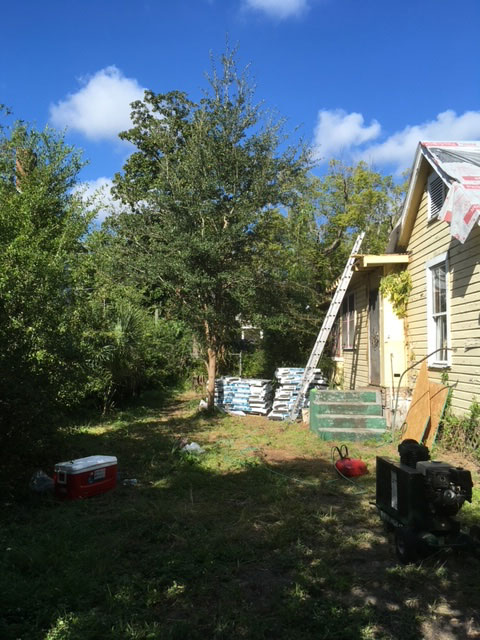 modern-farmhouse-during-side-yard-material-staging-jacksonville-mnm-homes-07-02 Renovation in progress: ladder against the house with stacked bagged materials in the overgrown side yard